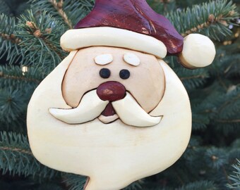 Puede incluir: Adorno navideño de madera de Papá Noel. El adorno presenta una cara de Papá Noel con un gorro burdeos, barba blanca y nariz rosada. El adorno cuelga de un hilo verde sobre un fondo de ramas de hoja perenne.