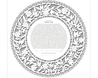 May include: A framed, circular papercut artwork with floral and leaf designs. The white papercut is layered over a grey background, creating a three-dimensional effect. The artwork contains Hebrew text within the circular design. The frame is white.
