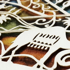 Op de afbeelding: Close-up van een wit, ingewikkeld uitgesneden papieren ontwerp met een vogel silhouet en bloemmotieven. De achtergrond toont een mix van kleuren, waaronder oranje, groen en geel, waardoor een glas-in-lood effect ontstaat. Het woord "peace" is zichtbaar.