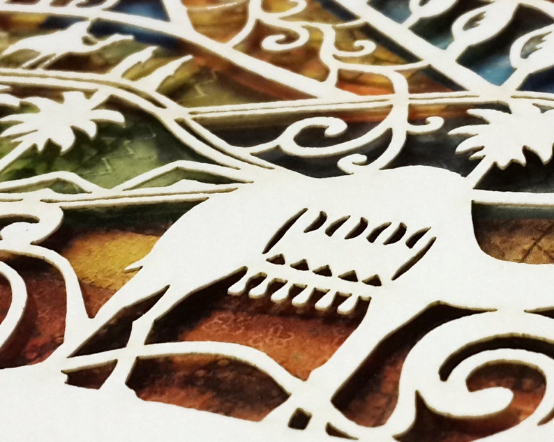 Op de afbeelding: Close-up van een wit, ingewikkeld uitgesneden papieren ontwerp met een vogel silhouet en bloemmotieven. De achtergrond toont een mix van kleuren, waaronder oranje, groen en geel, waardoor een glas-in-lood effect ontstaat. Het woord "peace" is zichtbaar.