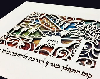 May include: White paper cut art with a desert scene featuring a camel, palm trees, and decorative scrollwork. The artwork has a colorful background and Hebrew text at the bottom. The paper is mounted on a white background.