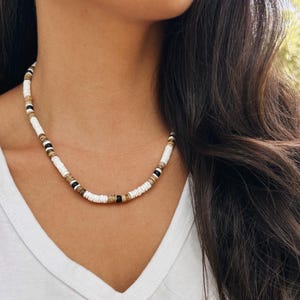 May include: A beaded necklace featuring a repeating pattern of small, flat, circular disc beads in white, tan, and black. The necklace is constructed from numerous thin, stacked discs that create a textured, cylindrical appearance around the neck.