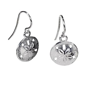May include: A pair of silver-colored drop earrings. Each earring features a circular, textured pendant with a sand dollar design. The earrings have a simple hook closure.