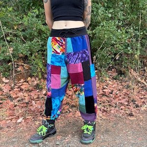 May include: Patchwork pants featuring a variety of colorful fabric squares in shades of blue, purple, pink, and green. The design includes tie-dye patterns, solid black panels, and textured fabric sections. The pants have a black elastic waistband with a drawstring.