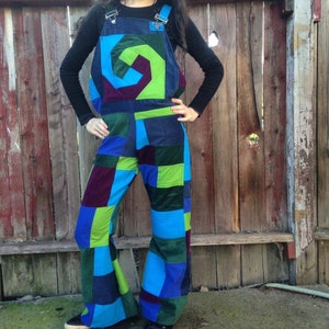 Op de afbeelding: Een overall met patchwork-ontwerp in tinten blauw, groen en paars. De slab heeft een spiraalvormig ontwerp. De overall heeft verstelbare bandjes en een wijde pijpen. De persoon draagt er een zwart shirt met lange mouwen onder.