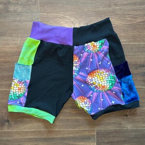 Op de afbeelding: Patchwork shorts met een discobal ontwerp. De shorts hebben panelen van zwart, paars, groen en blauw fluweel. De discobal print staat op een paarse achtergrond met stralende lijnen.