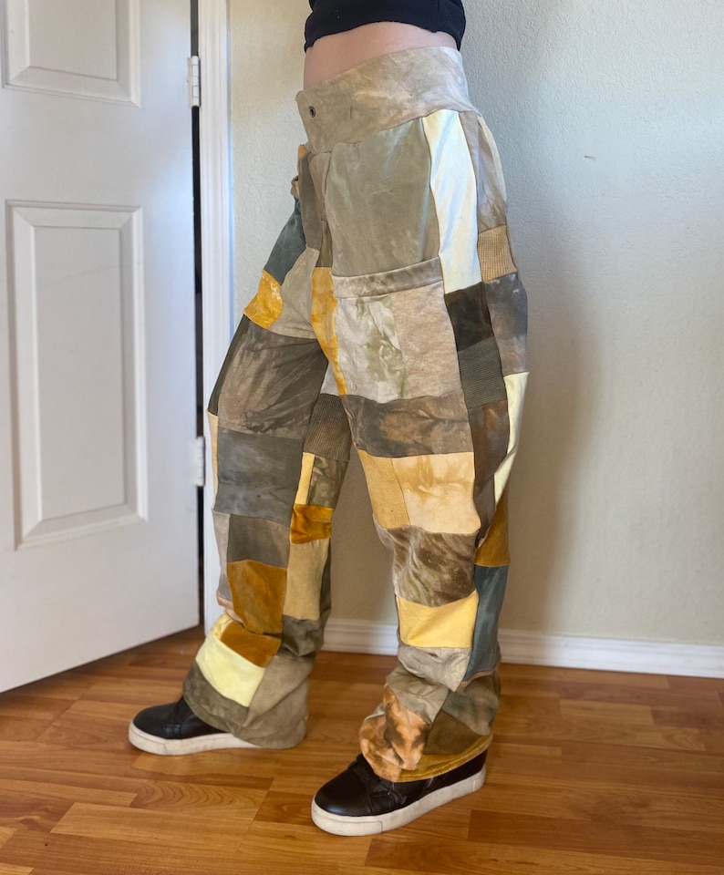 K&ouml;nnte beinhalten: Patchworkhose in den Farbt&ouml;nen Beige, Braun und Gelb. Die Hose hat eine hohe Taille und ein weites Bein. Der Stoff scheint eine Mischung aus Texturen zu sein, mit einer Vielzahl von quadratischen und rechteckigen Flicken, die zusammengen&auml;ht sind. Die Hose wird mit schwarzen Turnschuhen getragen.