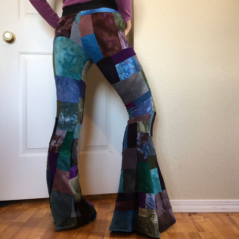 Op de afbeelding: Patchwork broek met uitlopende pijpen, bestaande uit diverse stoffen vierkanten in tinten blauw, paars, groen en zwart. De broek heeft een wijd uitlopend silhouet en een effen zwarte tailleband, opgebouwd uit meerdere textuurrijke stofpanelen.
