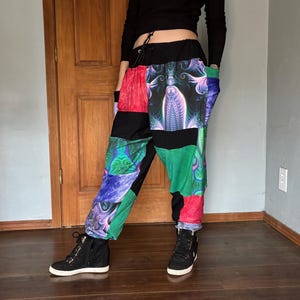 May include: Patchwork pants with a black waistband and drawstring. The pants feature panels of red, green, blue, and black fabric with a psychedelic design. The person is wearing black high-top sneakers.