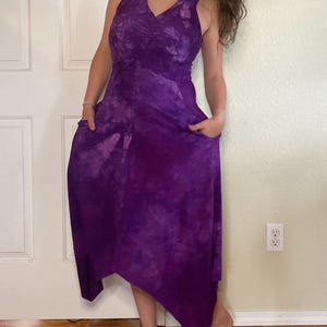 May include: A sleeveless, tie-dye purple dress with a V-neck and pockets. The dress has a flowing, asymmetrical hemline. The fabric has a marbled effect, with shades of deep purple and lighter lavender. The model is wearing the dress.