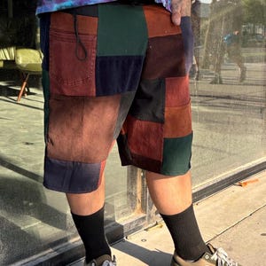 Peut inclure: Short en patchwork de velours côtelé dans les tons marron, vert et bleu marine. Le short a une taille à cordon et des poches. La personne porte des chaussettes noires et des baskets vertes et beiges.