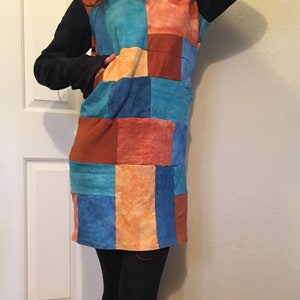 May include: A patchwork dress with a V-neck and long black sleeves. The dress features a colourful pattern of squares in shades of blue, orange, and red. The dress is knee-length and worn with black leggings.