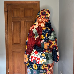 Puede incluir: Una manta con capucha con un diseño de patchwork con patrones florales y geométricos en tonos de rojo, azul, amarillo y verde. La manta parece estar hecha de un material suave y afelpado, que ofrece calidez y comodidad.