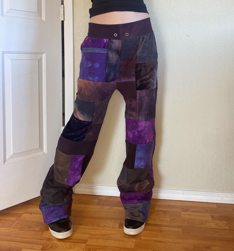 K&ouml;nnte beinhalten: Patchwork-Hose in Lila-, Braun- und Graut&ouml;nen. Die Hose hat einen hohen Bund mit zwei Metall&ouml;sen und eine lockere, bequeme Passform. Der Stoff scheint eine Mischung aus Samt und anderen Materialien zu sein, wodurch eine einzigartige Textur entsteht.