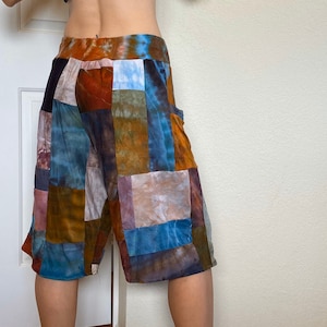 Puede incluir: Pantalones cortos de patchwork con un estampado tie-dye en tonos azules, marrones y beige. Los pantalones cortos tienen una cintura alta y un corte holgado.