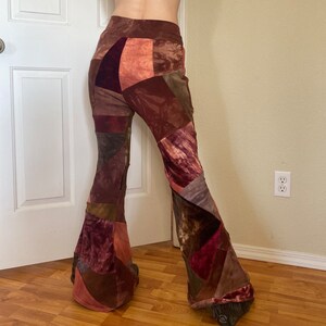 Op de afbeelding: High-waisted broek met uitlopende pijpen en een patchwork-ontwerp in diverse tinten bruin, bordeaux en roest. Het kledingstuk is gemaakt van verschillende stoftexturen, waaronder fluweel en corduroy, gerangschikt in een asymmetrisch geometrisch patroon.