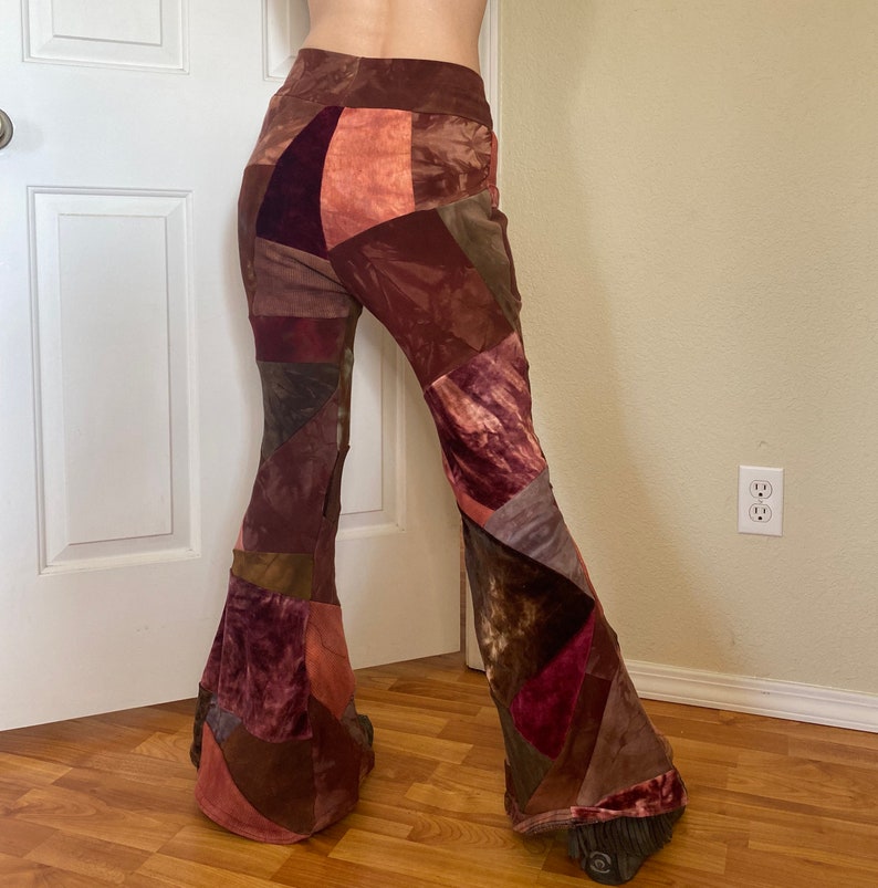 Op de afbeelding: High-waisted broek met uitlopende pijpen en een patchwork-ontwerp in diverse tinten bruin, bordeaux en roest. Het kledingstuk is gemaakt van verschillende stoftexturen, waaronder fluweel en corduroy, gerangschikt in een asymmetrisch geometrisch patroon.