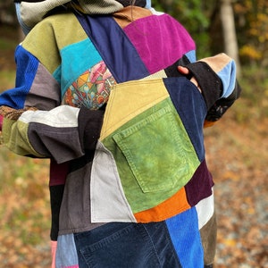 May include: A patchwork hoodie with a variety of corduroy patches in shades of blue, green, purple, and yellow. The hoodie features a front pocket and a hood with a striped pattern. The design includes a colorful mushroom graphic.