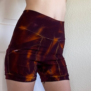 May include: High-waisted, tie-dye shorts in shades of burgundy and orange. The shorts have a fitted design with a high waistband and a short leg length. The fabric has a marbled, tie-dye pattern with visible seams.
