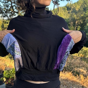 May include: A black long-sleeve mock neck top featuring two large front pockets. The pockets are constructed from a mix of light blue fabric, purple velvet, and a blue patterned textile with a geometric star design, finished with a black hem.