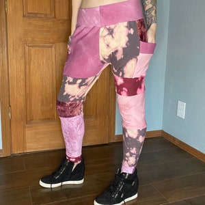 May include: Patchwork pants in shades of pink and burgundy with a tie-dye effect. The pants have a wide waistband and pockets. The model is wearing black high-top sneakers with white soles.