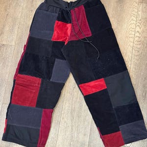 May include: Patchwork pants in a wide-leg style, featuring a mix of black, red, and gray fabrics. The pants have a drawstring waist and a variety of textures, including corduroy and velvet. A unique and stylish garment.