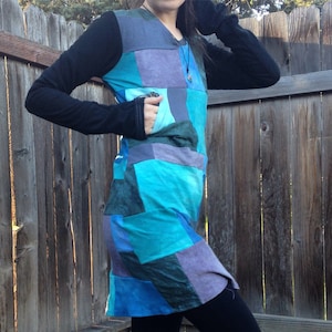 Könnte beinhalten: Ein Patchwork-Tunika-Kleid in Blau-, Türkis- und Lilatönen. Das Kleid hat lange schwarze Ärmel und einen V-Ausschnitt. Das Design integriert rechteckige Stoffflicken und schafft einen einzigartigen, künstlerischen Look. Das Kleid hat eine Tasche auf der Vorderseite.