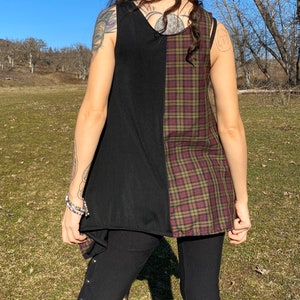 May include: A sleeveless top with a unique design, featuring a split construction. One side is solid black, while the other showcases a plaid pattern in shades of burgundy, olive green, and gold. The top is worn with black leggings.