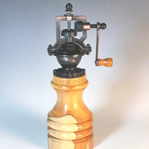 May include: A wooden pepper mill with a light brown, turned wood base and a dark metal grinding mechanism. The mill features a wooden handle and a decorative top. The wood grain is visible on the base.