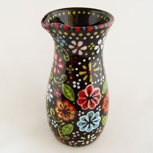 Puede incluir: Un jarrón de cerámica negro con un patrón floral. El jarrón está decorado con flores coloridas, hojas y puntos. El jarrón tiene una boca ancha y una base estrecha.