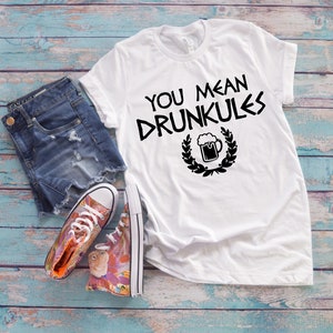 May include: White t-shirt with the text "YOU MEAN DRUNKULES" in black, along with a beer mug graphic. The shirt is paired with denim shorts and colorful sneakers on a blue and white wooden surface.