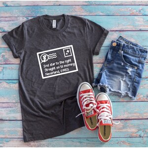 Op de afbeelding: Een donkergrijs t-shirt met een witte afbeelding van een postzegel met de tekst "2nd star to the right, Straight on til morning, Neverland, 01953." De outfit wordt gecompleteerd met een spijkerbroekshort en rode sneakers.