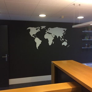 Puede incluir: Un contorno blanco de un mapa del mundo en una pared negra en un entorno de oficina. El mapa est&aacute; en la pared detr&aacute;s de una mesa de madera y un estante con plantas en macetas.
