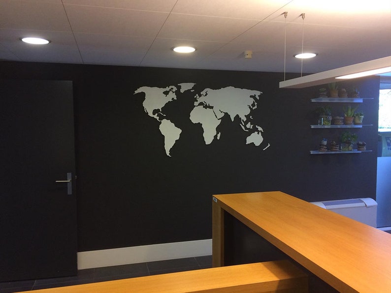 Puede incluir: Un contorno blanco de un mapa del mundo en una pared negra en un entorno de oficina. El mapa est&aacute; en la pared detr&aacute;s de una mesa de madera y un estante con plantas en macetas.
