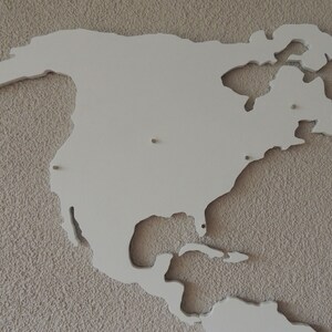 Puede incluir: Un recorte de madera blanco de Am&eacute;rica del Norte, montado en una pared beige texturizada. El recorte es una representaci&oacute;n simplificada del continente, con las principales masas terrestres y algunas de las islas circundantes.