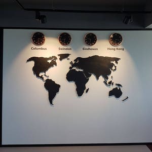 Puede incluir: Un mapa del mundo en blanco y negro con cuatro relojes en una pared blanca. Los relojes est&aacute;n etiquetados como "Columbus", "Swindon", "Eindhoven" y "Hong Kong".