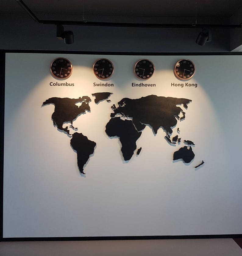 Puede incluir: Un mapa del mundo en blanco y negro con cuatro relojes en una pared blanca. Los relojes est&aacute;n etiquetados como "Columbus", "Swindon", "Eindhoven" y "Hong Kong".
