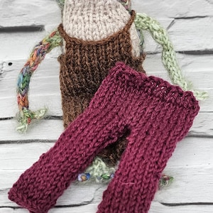 Puede incluir: Una muñeca de rana de punto que lleva un mono marrón y pantalones granate. La rana tiene un cuerpo de punto colorido, multicolor y ojos grandes y negros.