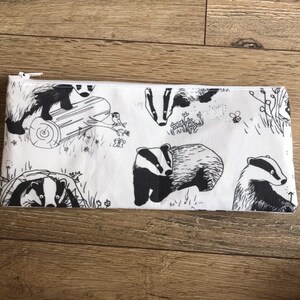 May include: A white zippered pouch with a black and white print of badgers, leaves, and a bee. The badgers are in various poses, some are walking, some are standing, and some are looking at the viewer.