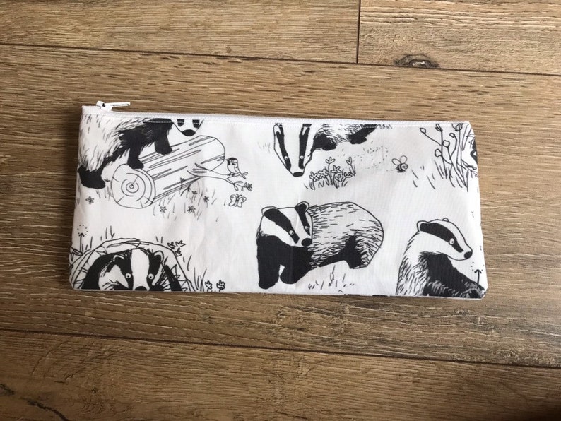 May include: A white zippered pouch with a black and white print of badgers, leaves, and a bee. The badgers are in various poses, some are walking, some are standing, and some are looking at the viewer.