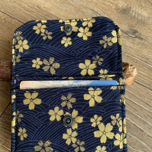 May include: Open navy blue fabric wallet with a gold floral pattern. The wallet is open, revealing a card inside. The design features a repeating pattern of gold flowers and wave-like lines. The wallet is secured with a snap closure.