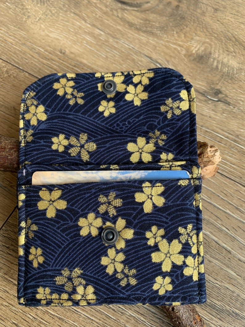 May include: Open navy blue fabric wallet with a gold floral pattern. The wallet is open, revealing a card inside. The design features a repeating pattern of gold flowers and wave-like lines. The wallet is secured with a snap closure.