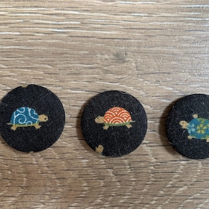 May include: Three black, circular fabric buttons with turtle designs. Each button features a different turtle design in shades of blue, orange, and yellow. The buttons are arranged on a wooden surface.