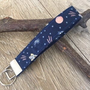 Peut inclure: Un porte-clés en tissu bleu avec un motif de fusée, d'étoiles et d'un cercle rose. Le porte-clés a un anneau en métal argenté.