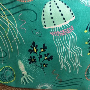 May include: Teal fabric with a repeating pattern of jellyfish, seaweed, and other sea life. The jellyfish are light green with pink and white accents. The seaweed is dark green and yellow.