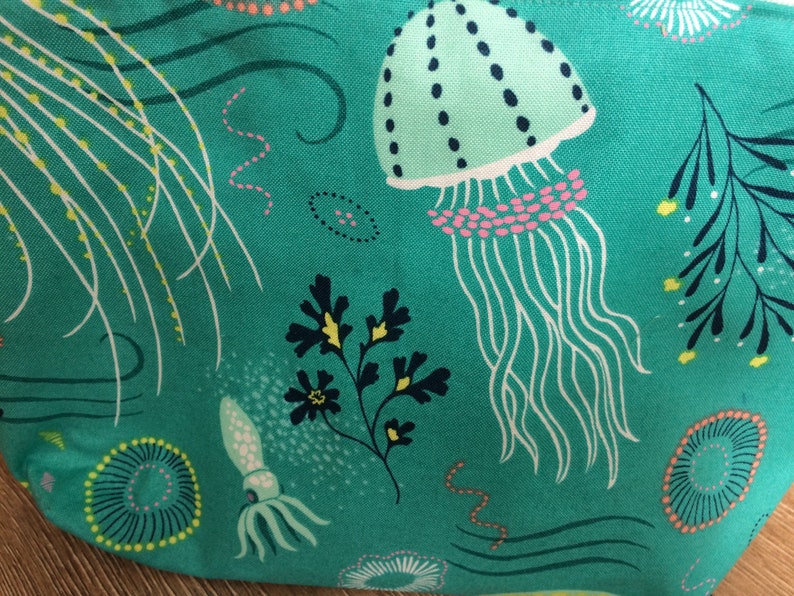 May include: Teal fabric with a repeating pattern of jellyfish, seaweed, and other sea life. The jellyfish are light green with pink and white accents. The seaweed is dark green and yellow.