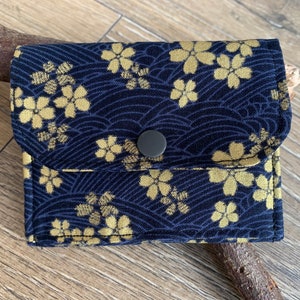 May include: A small, navy blue wallet with a gold floral pattern and a wave design. The wallet has a flap closure with a gray button. The fabric has a textured appearance. The wallet is sitting on a wooden surface.