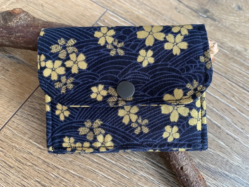 May include: A small, navy blue wallet with a gold floral pattern and a wave design. The wallet has a flap closure with a gray button. The fabric has a textured appearance. The wallet is sitting on a wooden surface.