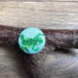 May include: A light blue button with a green frog design. The frog is facing right and has a spotted pattern.