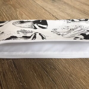 May include: A white zippered pouch with a black and white print of badgers. The pouch is lying flat on a wooden surface.